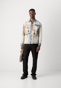 Bunt gemusterte Shirtjacke mit einem hellbeigen Körper, blauen Kragen, Taschen und bedruckten Designs. Schwarze Hosen und schwarze Schuhe runden den Look ab.