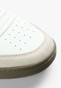 Acercamiento al dedo de una zapatilla blanca con parte superior de cuero perforado, ribete de ante beige y suela de goma texturizada.