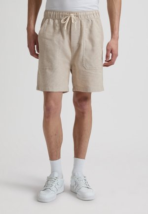 Mann trägt beige Shorts mit Kordelzug und aufgesetzten Taschen vorne, weiße Kniestrümpfe und weiße Turnschuhe, steht vor einem einfarbigen Hintergrund.