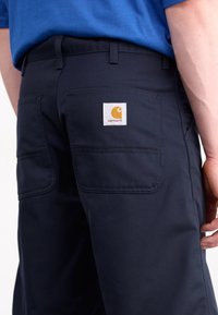 Navy blue work pants with two back pockets and a square white patch featuring an orange logo. Made from durable fabric with a smooth texture.