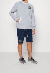 Sudadera gris claro con cuello de canalé y bolsillo frontal, combinada con pantalones cortos azul marino que tienen bolsillos laterales y un logotipo de parche. Zapatillas grises.