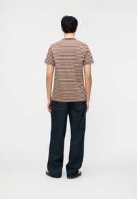 Άνδρας που φοράει καφέ και λευκή ριγέ κοντομάνικη μπλούζα και σκούρο μπλε παντελόνι με φαρδιά μπατζάκια, στέκεται με την πλάτη στραμμένη σε απλό ανοιχτόχρωμο φόντο.