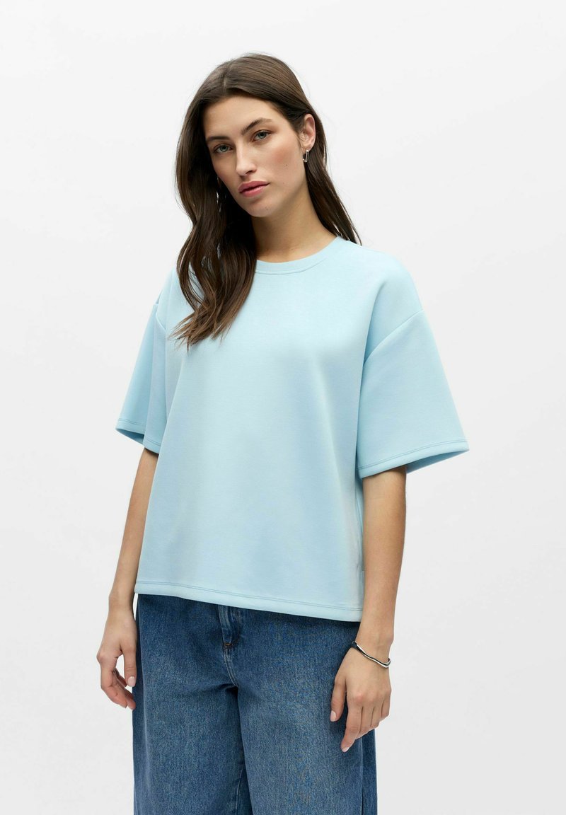 T-shirt oversize bleu clair à manches courtes, col rond, texture lisse, associé à un jean décontracté ample.