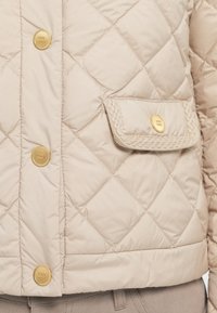 Beige quilted jacket featuring diamond pattern, gold-tone buttons, and a braided pocket flap. Smooth texture with a fitted silhouette.