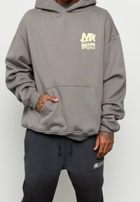 Szary hoodie z kieszenią z przodu, ozdobiony żółtym graficznym nadrukiem na klatce piersiowej. Ma luźny krój oraz ściągacze na mankietach, w połączeniu z ciemnymi spodniami.