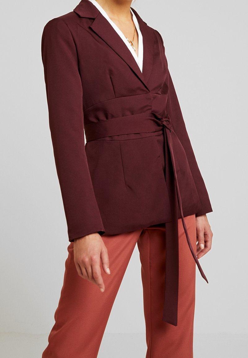 Femme portant un blazer bordeaux foncé ceinturé sur un haut blanc et un pantalon couleur rouille, sur un fond gris clair uni.