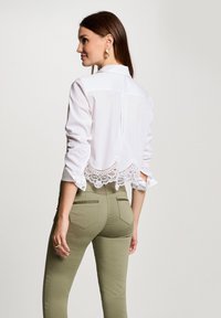 Weißes Shirt mit Spitze am Saum und hochgekrempelten Ärmeln, kombiniert mit olivgrünen, taillierten Hosen mit Gesäßtaschen und Details.