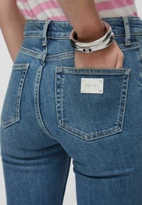 Jean en denim bleu taille haute, avec deux poches arrière, dont une avec une étiquette de marque blanche rectangulaire. Texture lisse et coupe ajustée.
