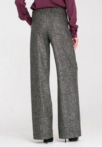 Kobieta ubrana w szare, szerokie spodnie z wysokim stanem oraz fioletową bluzkę z długim rękawem, stojąca z rękami wzdłuż ciała, pokazująca widok z tyłu.