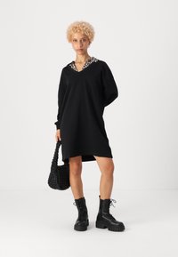 Črna oversized obleka z ovratnikom v leopardjem vzorcu, z dolgimi rokavi in ohlapnim krojem. V družbi debelih črnih čevljev z vezalkami in tkano torbo.