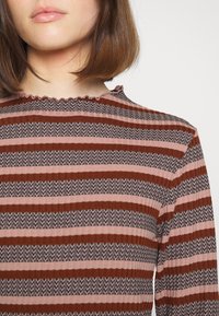 Pruhovaný svetr v teplých tónech hnědé a růžové. Má texturovaný vzor a zvlněný výstřih. Materiál vypadá jako vroubkovaný a vypasovaný.