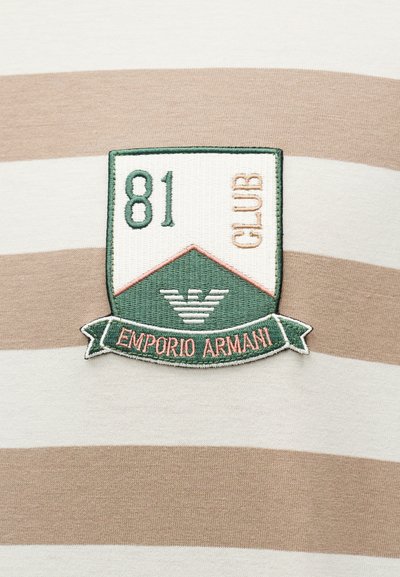 Υφασμάτινο έμπλαστρο σε σχήμα ασπίδας πάνω σε μπεζ και λευκό υφαντό με ρίγες, με κεντημένα τα λόγια "81 CLUB" και "Emporio Armani" σε πράσινους και ροδακινί τόνους.