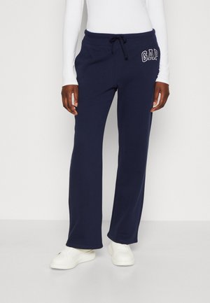 Vrouw die marineblauwe GAP joggingbroek met trekkoord draagt en witte sneakers, staand tegen een effen lichte achtergrond.