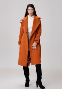 Long manteau orange en tissu épais avec un large col, associé à un pull col roulé blanc et une jupe grise. Des bottes noires montantes complètent le look.