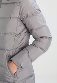 Grijze donzen jas met horizontale doorgestikte lijnen, voorzien van een zijzak met ritssluiting en een logopatch op de schouder. Glad, glanzend oppervlak.