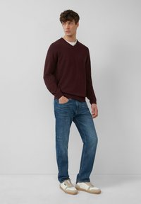 Mann trägt einen burgunderfarbenen V-Ausschnitt-Pullover, ein weißes Unterhemd, blaue Jeans und weiße Sneaker und steht mit einer Hand in der Tasche.
