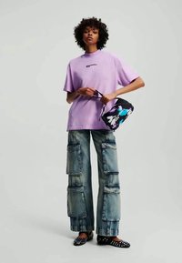 Lilla oversized t-shirt med logo, slidte blå cargo bukser med lommer, og sorte loafers med hvide prikker. Lille grafisk taske i hånden.