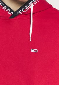 Sweat à capuche rouge avec cordon blanc et petit logo rectangulaire en rouge, blanc et bleu sur la poitrine, doublure intérieure de la capuche noire avec texte blanc.