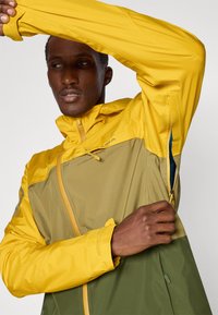 Gelbe und olivgrüne Jacke mit Reißverschluss vorne, verstellbaren Bündchen und Seitentasche, bestehend aus einem glatten Material und leichtem Stoff.