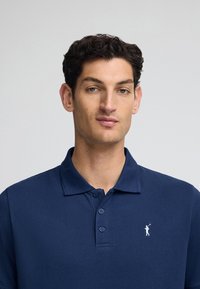 Marinblå pikétröja i texturerad tyg, med en tvåknappsknäppning och en liten vit logotyp på vänster bröst.