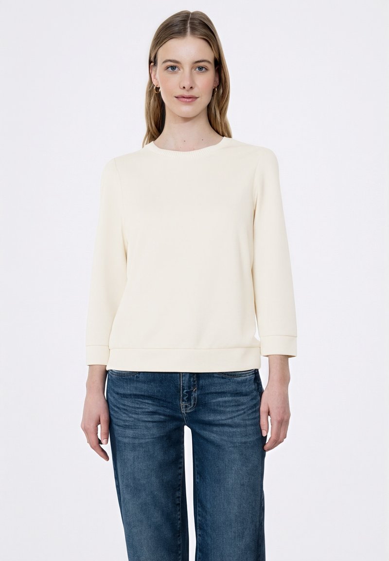 Jasnokremowa bluza z okrągłym dekoltem, rękawami o długości do łokcia, ściągaczami na mankietach oraz prostym dołem, zestawiona z ciemnoniebieskimi dżinsami.