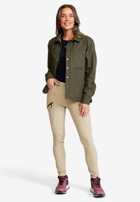 Kvinna står klädd i en olivgrön knappad jacka, svart topp, beige smala byxor med blixtlås-ficka, och rosa vandringsskor, leende med handen i fickan.