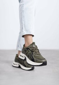 Zapatillas deportivas en verde oliva, confeccionadas con material de malla y sintético, con una suela blanca gruesa y detalles en negro, diseñadas para ofrecer comodidad y soporte.
