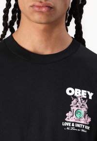 Svart bomull t-skjorte med grafisk trykk i rosa og grønt, med teksten "OBEY LOVE & UNITY NOW" foran. Korte ermer, rund hals.