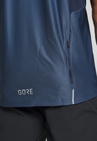 Námornícké sportovní tričko s texturovaným povrchem, vybavené boční kapsou na zip a brandingem GORE. Hladká tkanina a elegantní designové prvky.