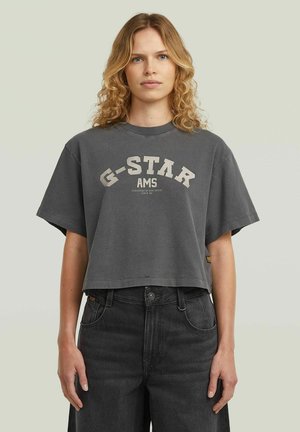 Młoda kobieta z kręconymi blond włosami, ubrana w ciemnoszary krótki T-shirt z napisem "G-STAR AMS" i czarne jeansy z wysokim stanem.