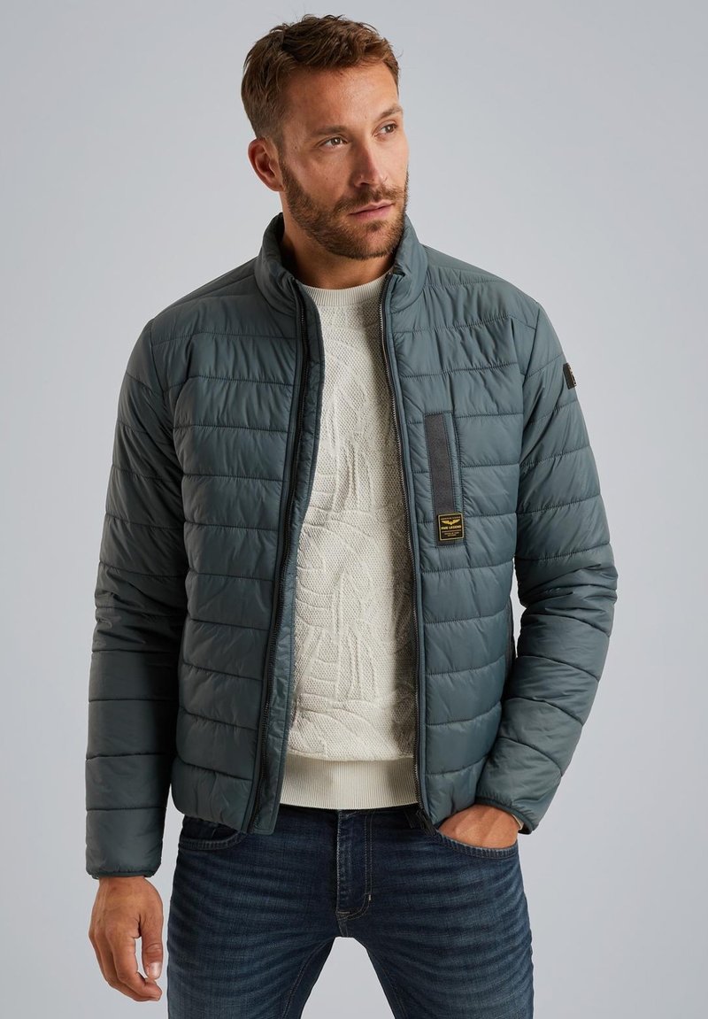 Giacca imbottita in verde scuro, con colletto alto, zip frontale e un maglione in maglia testurizzata sotto. Include dettagli decorativi in metallo.
