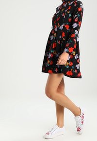 Schwarzes Blumenkleid mit langen Ärmeln, verziert mit roten und lila Blumen. Kombiniert mit weißen Sneakern, die mit floralem Stickmuster geschmückt sind.