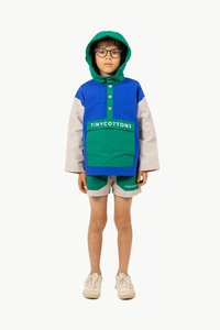 Veste colorée en bleu et vert, avec une capuche, des boutons et une poche avant. Associée à un short clair arborant le logo de la marque.