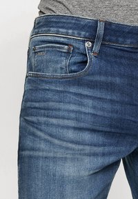 Blå denimjeans med en lätt blekning, utrustade med en framficka, metallknapp och sömmar längs kanterna.