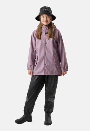 Giovane che indossa una giacca antipioggia viola, pantaloni impermeabili neri, stivali da pioggia neri e un cappello a secchiello nero, in piedi davanti a uno sfondo bianco.