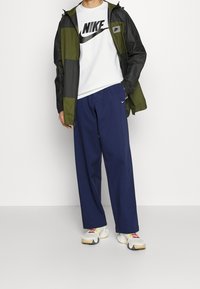 Nike vindjacka i grönt och svart, vit t-shirt med grafik, marinblå löst sittande byxor och mångfärgade sneakers. Slätt tyg, atletisk design.