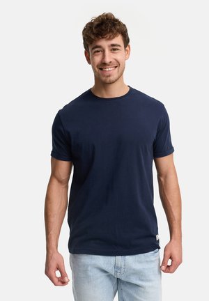 Ung mand med kort krøllet hår iført en mørkeblå t-shirt og lyseblå jeans, smilende og stående foran en hvid baggrund.
