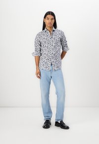 Hvit skjorte med blå blomstermønster, knapp-ned krage, og rullede ermer, kombinert med lyseblå jeans og svarte sko.
