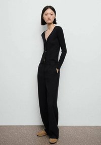 Svart ribbad cardigan med djup V-ringning, försedd med framknappar, ihop med lösa svarta byxor och ljusbruna platta skor.