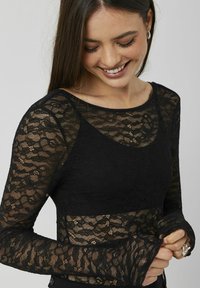 Haut en dentelle noire avec des manches longues, un col rond et un buste ajusté. Le tissu présente des motifs floraux délicats et une texture transparente.