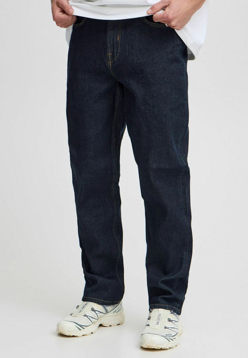 Dark blue denim jeans with a straight-leg cut, featuring visible stitching and a classic five-pocket design, paired with white sneakers.