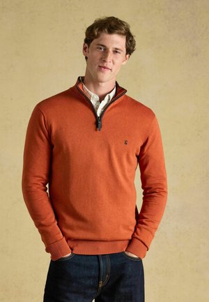 Jonge man met een oranje trui met rits en een wit geruit overhemd, met handen in de zakken, staand tegen een effen achtergrond.