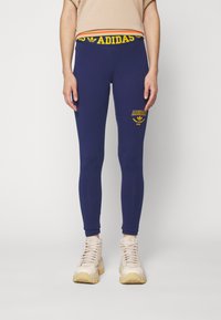 Mornarske modre legice z elastičnim pasom Adidas, ki vsebujejo rumeno besedilo logotipa in majhen simbol na stegnu. Gladko blago, prilegan kroj.