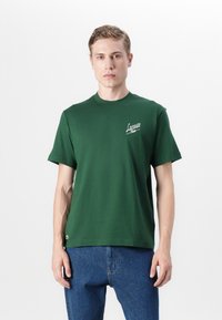 T-shirt en coton vert avec un col rond, des manches courtes et un logo Lacoste blanc sur la poitrine gauche. Porté avec un jean en denim.