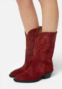 Botas de vaquero de ante rojo con punta afilada, patrones de costura intrincados y un tacón corto y cuadrado. El eje presenta un diseño de parte superior con formas scalloped.