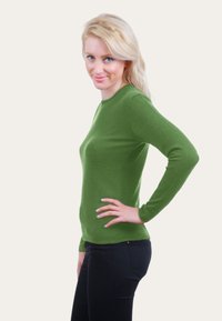 Femme blonde se tenant de profil avec un léger sourire, portant un haut à manches longues vert et un pantalon noir, la main reposant sur sa hanche.