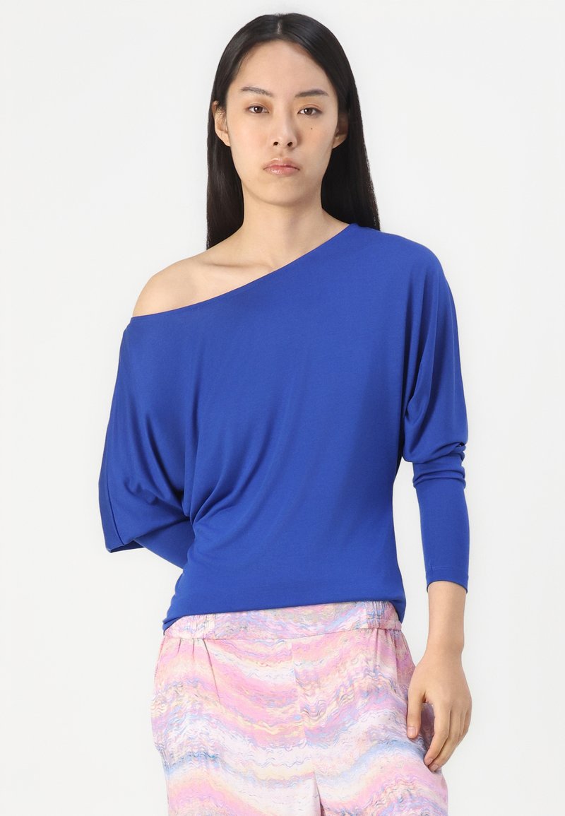 Blaues Off-Shoulder-Oberteil aus weichem Stoff mit langen Ärmeln; das drapierte Design betont die fließende Silhouette, kombiniert mit pinkfarbenen gemusterten Hosen.