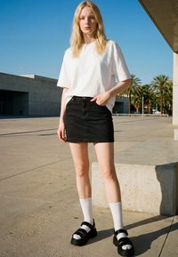 Jeune femme debout à l'extérieur portant un t-shirt blanc oversize, une jupe en denim noire, des chaussettes blanches et des sandales à plateformes noires.