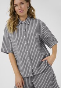 Chemise à manches courtes à rayures grises et blanches avec col, offrant une coupe décontractée et une texture douce, portée avec un pantalon assorti.