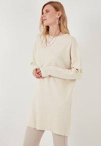 Pull en maille côtelée oversize de couleur crème avec des manches longues et des fentes latérales, doté d'un col rond et d'une texture subtile.